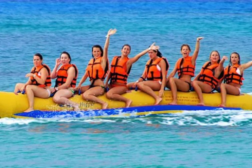 Eight girls in orange life jackets sitting on a banana boat (a long floating raft shaped like a banana)