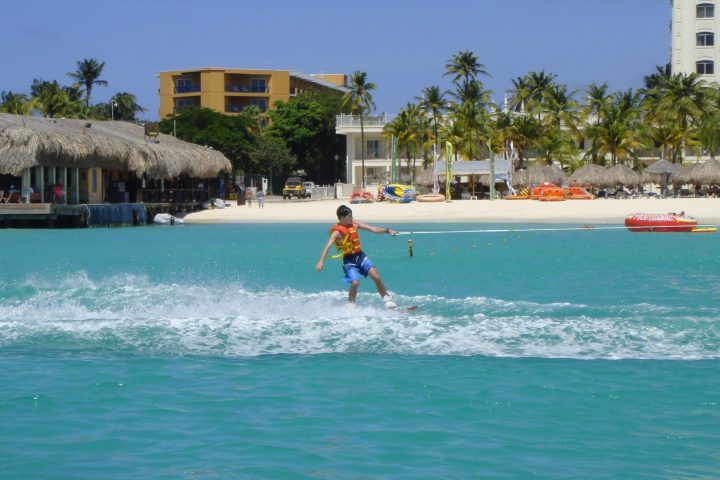 Boy in orange life jacket & blue swim trunks wakingboarding with hotels & cabanas on the beach are in the background.